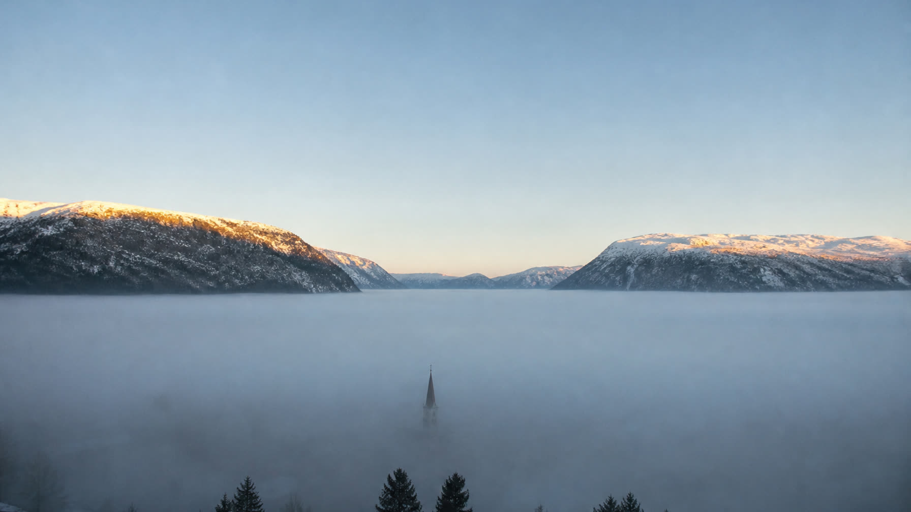 Norsk vinterdal fylt av kald tåke under klar himmel, med sol på fjelltoppene